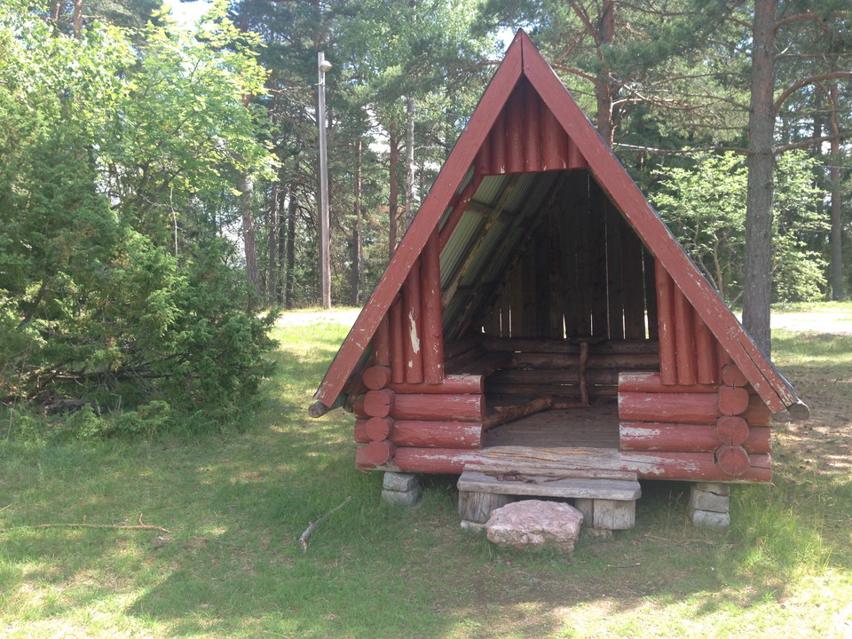 I en skogsglänta står ett vindskydd med högt i tak. Framför ingången finns en sten och en trappa som leder in i vindskyddet.
