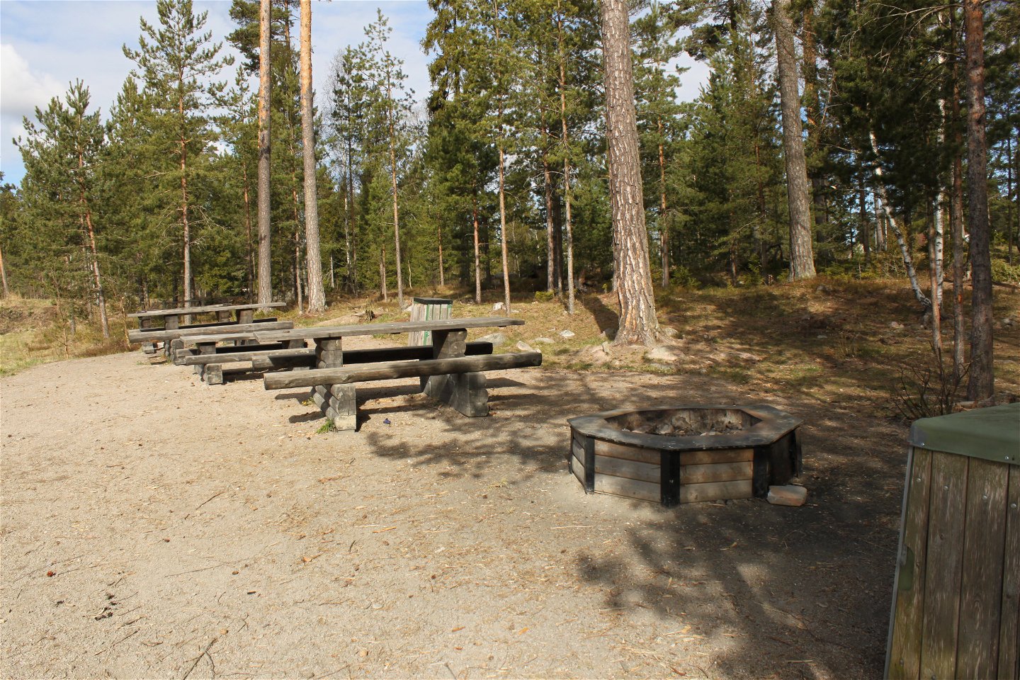 En grillplats står på jämn grusad yta. Vid grillplatsen finns flera bänkbord och soptunnor.