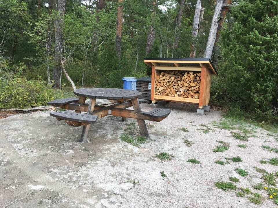Närbild på ett bänkbord, ett litet vedförråd och en soptunna som står i utkanten av en parkering.