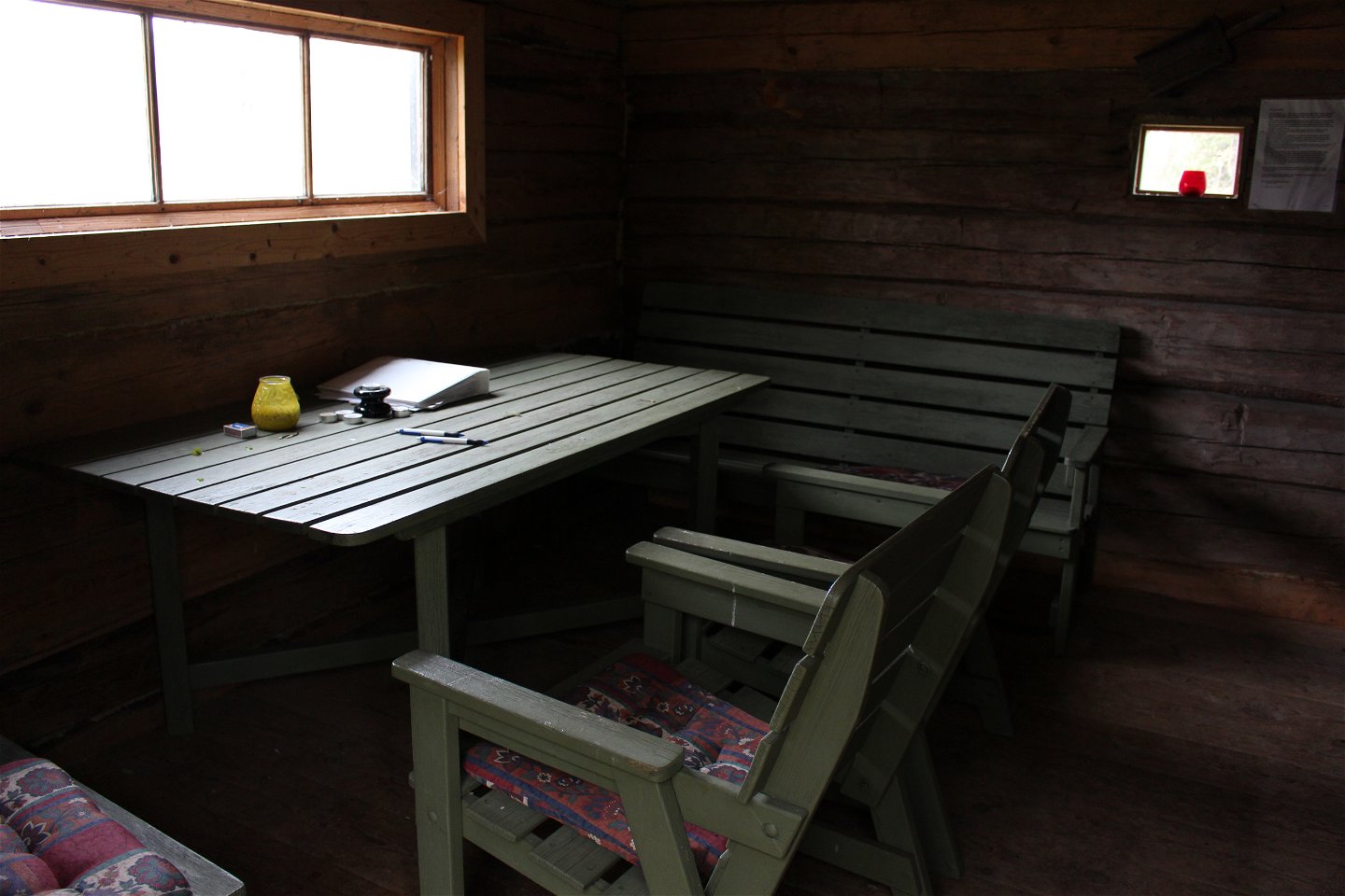 Vid ett smalt fönster står ett bord, två stolar och sittbänkar. På bordet finns pennor, värmeljus och en pärm.
