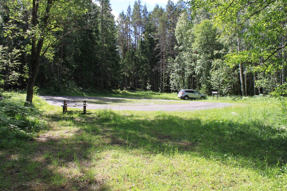 En bil står på en grusad parkeringsplats med skog runtom. I närheten står ett bänkbord.