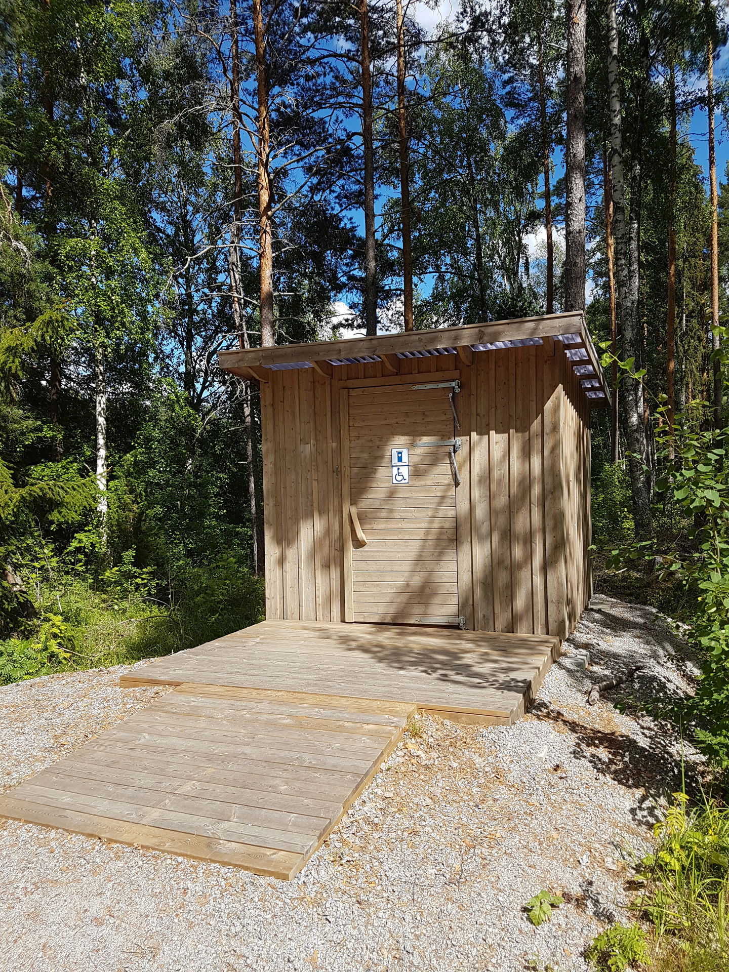 En ramp leder upp till en liten träplattform framför en tillgänglighetsanpassad torrtoalett. I bakgrunden är det skog.