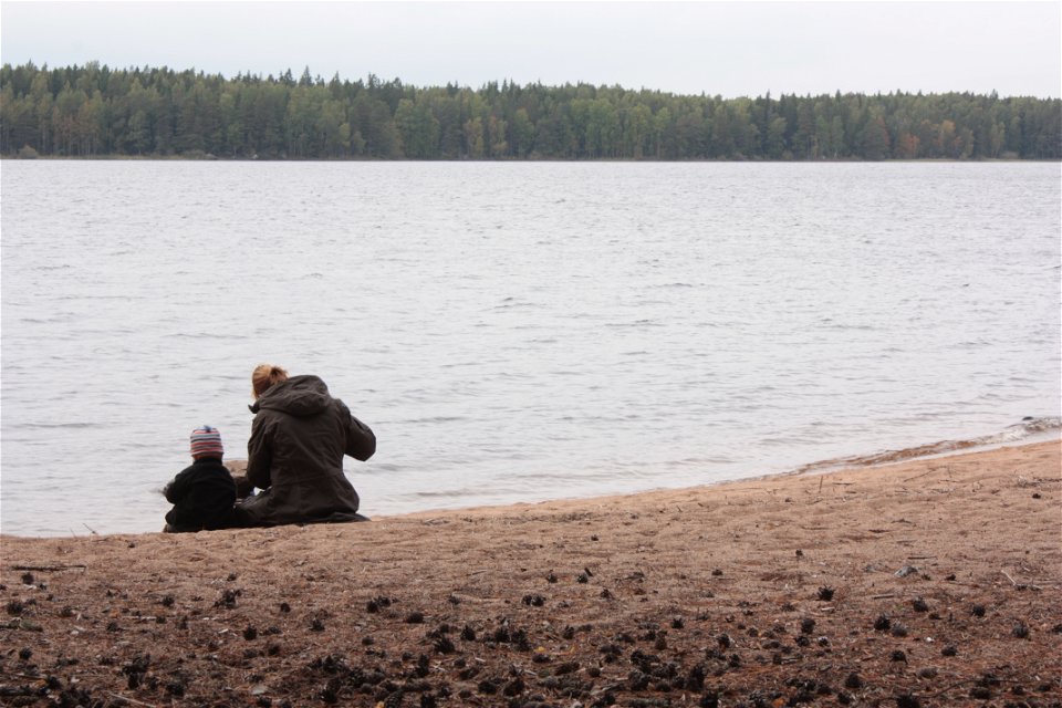 En vuxen och ett barn sitter bredvid varandra på en sandstrand vid vattnet