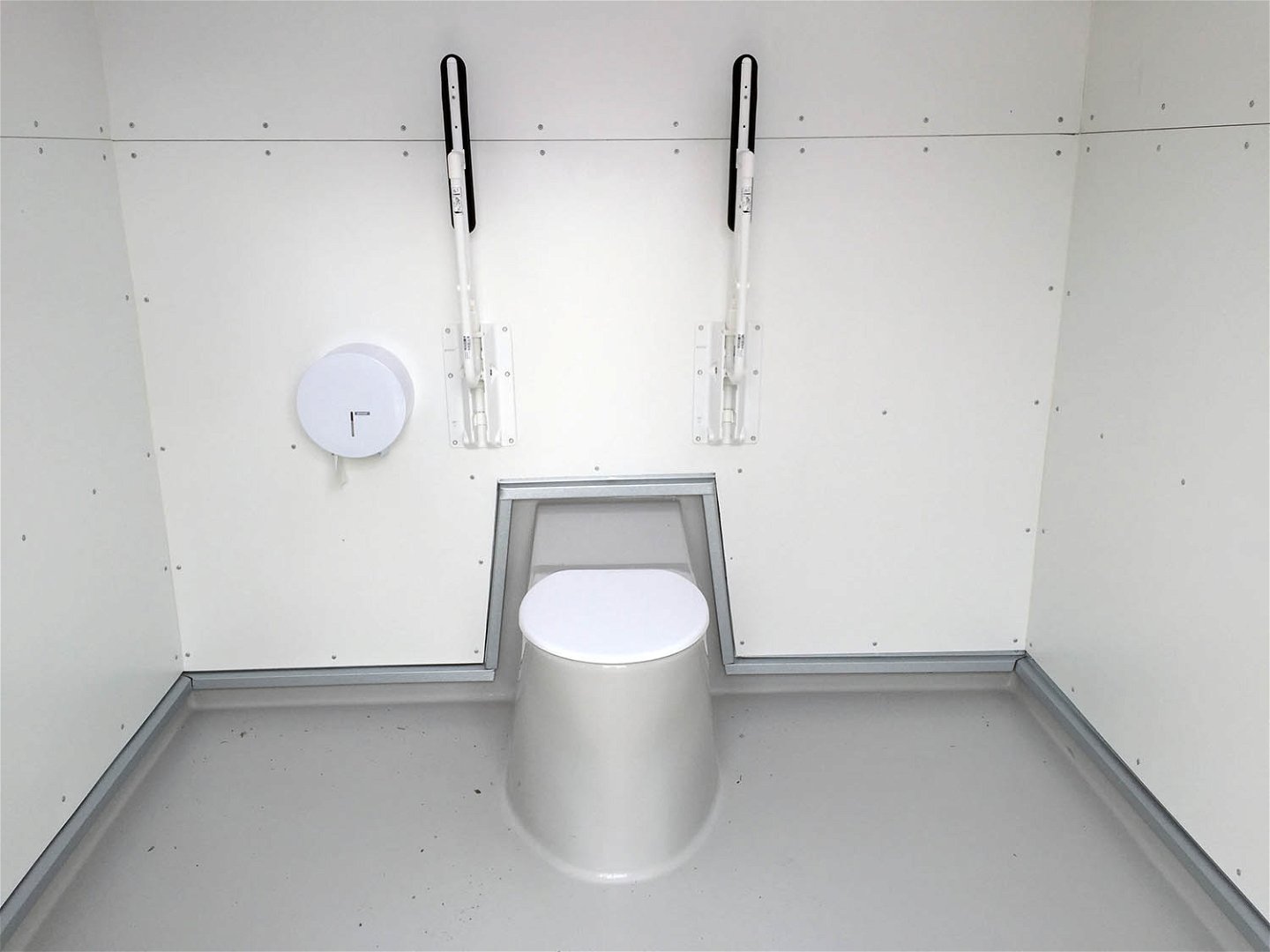 Toalettstol med locket stängt. Två armstöd uppfällda mot väggen. Toalettpappershållare till vänster om toalettstolen.