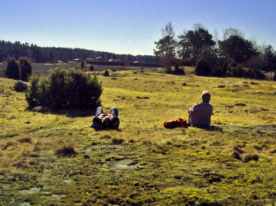 Två personer rastar på en vidsträckt hagmark. Ena personen sitter och den andra ligger på rygg och njuter av solen.