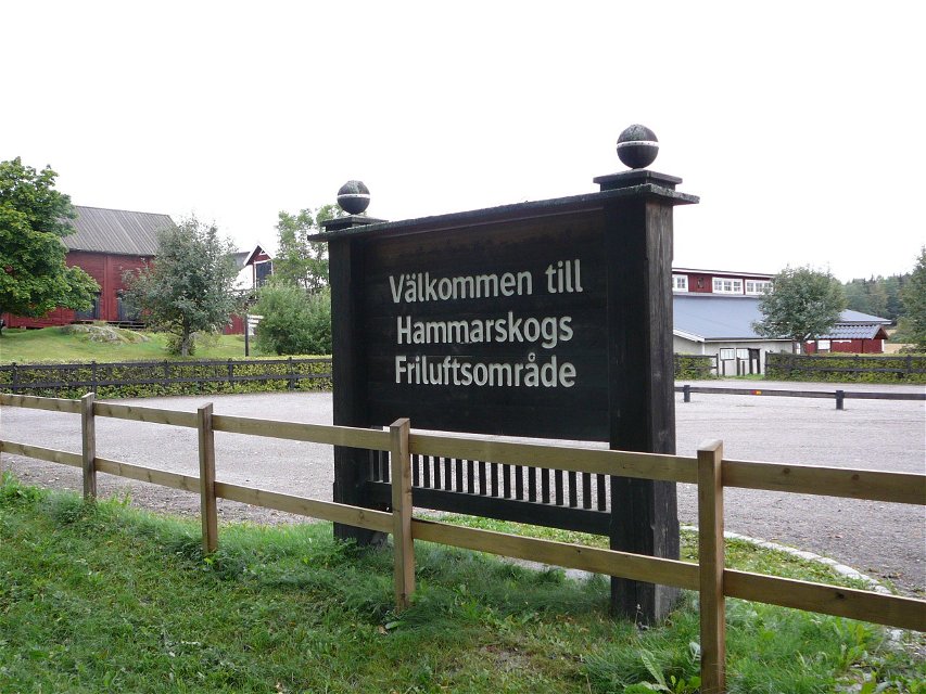 En stor skylt med texten "Välkommen till Hammarskogs friluftsområde" står vid en stor grusad parkeringsplats. Parkeringen är omgiven av ett staket.