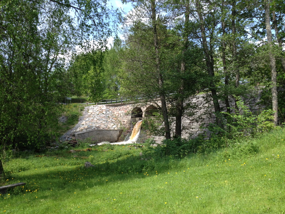 Under en stenbron forsar en å fram. Runtom finns skog och en öppen gräsyta. 