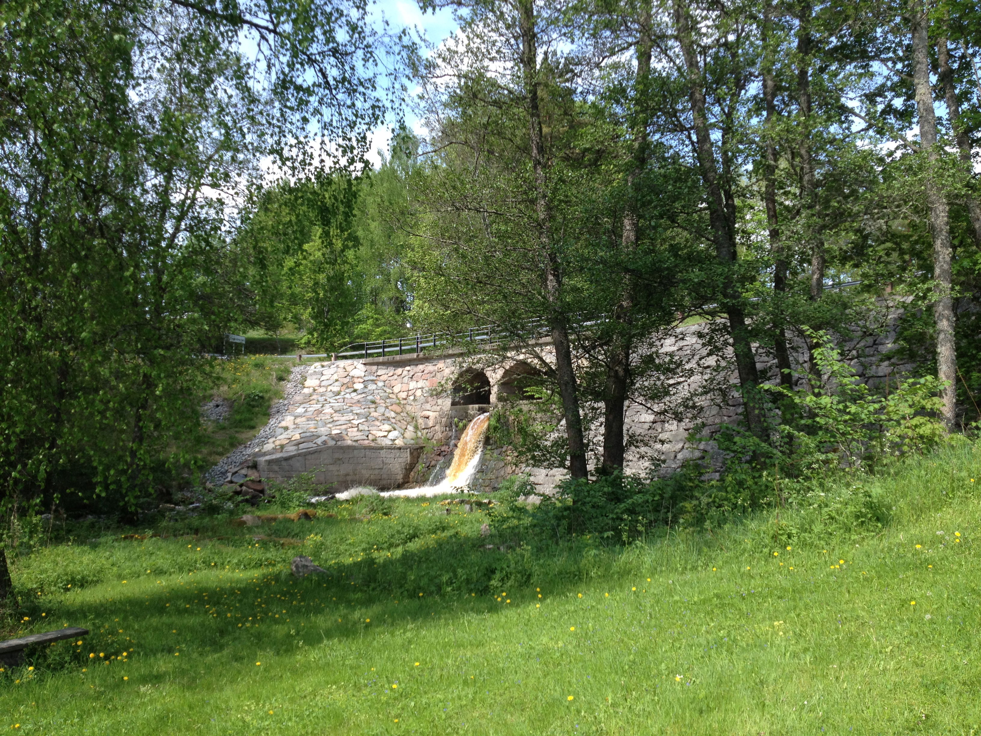 Under en stenbron forsar en å fram. Runtom finns skog och en öppen gräsyta. 