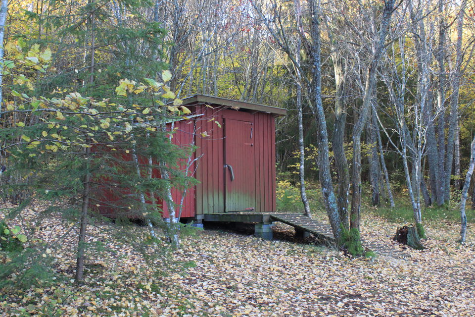 En torrtoalett står bland träd i naturen. En träramp leder upp till toalettdörren.