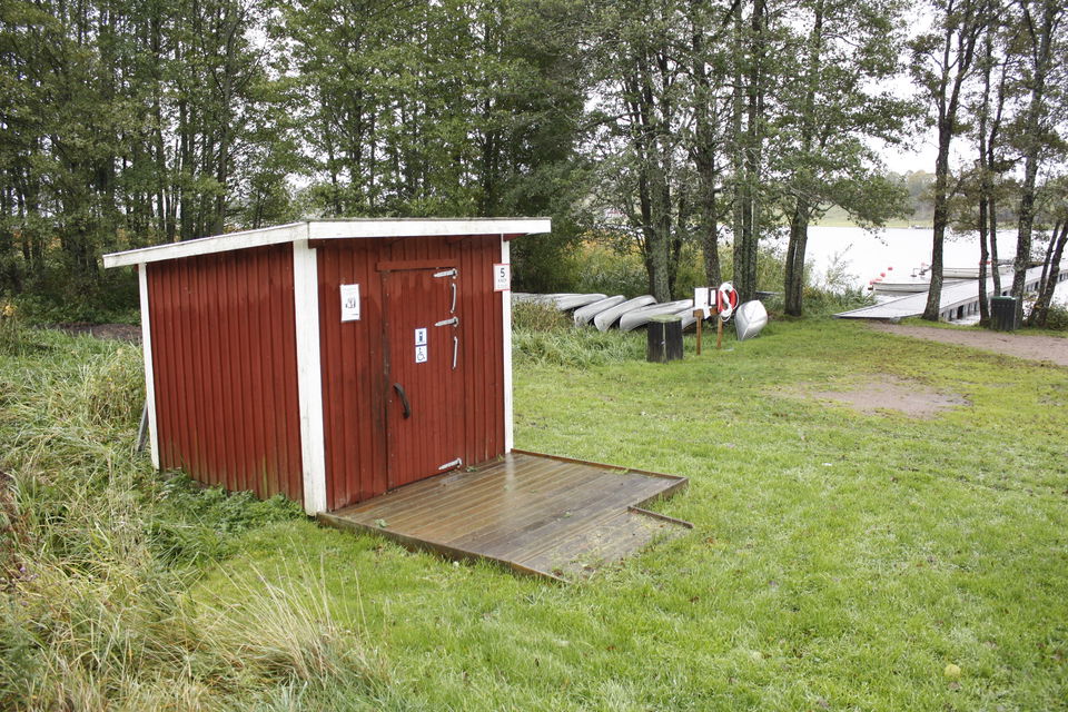 En bred ramp leder upp till en tillgänglighetsanpassad torrtoalett. I bakgrunden finns vatten och flera kanoter. 