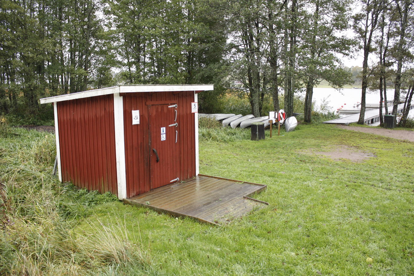 En bred ramp leder upp till en tillgänglighetsanpassad torrtoalett. I bakgrunden finns vatten och flera kanoter. 
