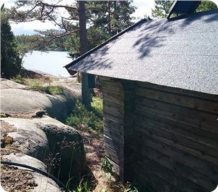 Fotot är taget uppe på ett klibblock. Vid klippblocket står en bastu och i bakgrunden syns havet mellan trädstammarna.