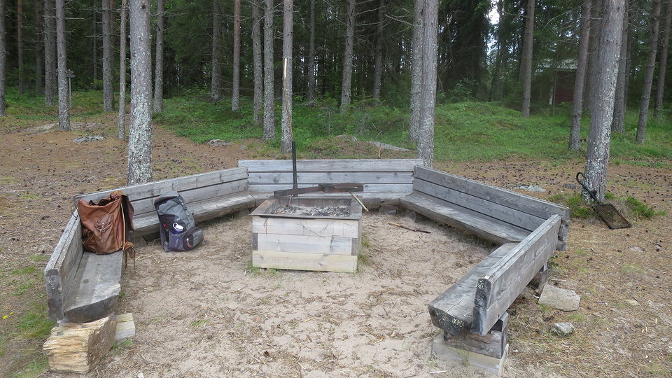 Eldstad i sand omgiven av bänkar på tre sidor och skog bakom.