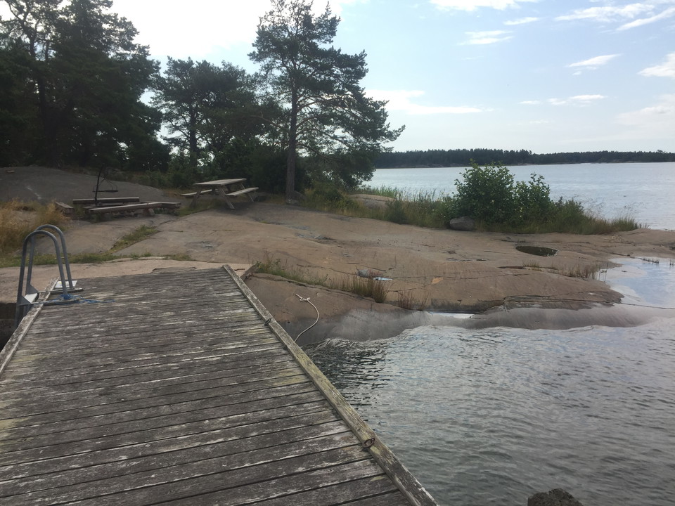Från badbryggan ser man en grillplats och en sittbänk som står på en sluttande stenhäll vid vattnet.