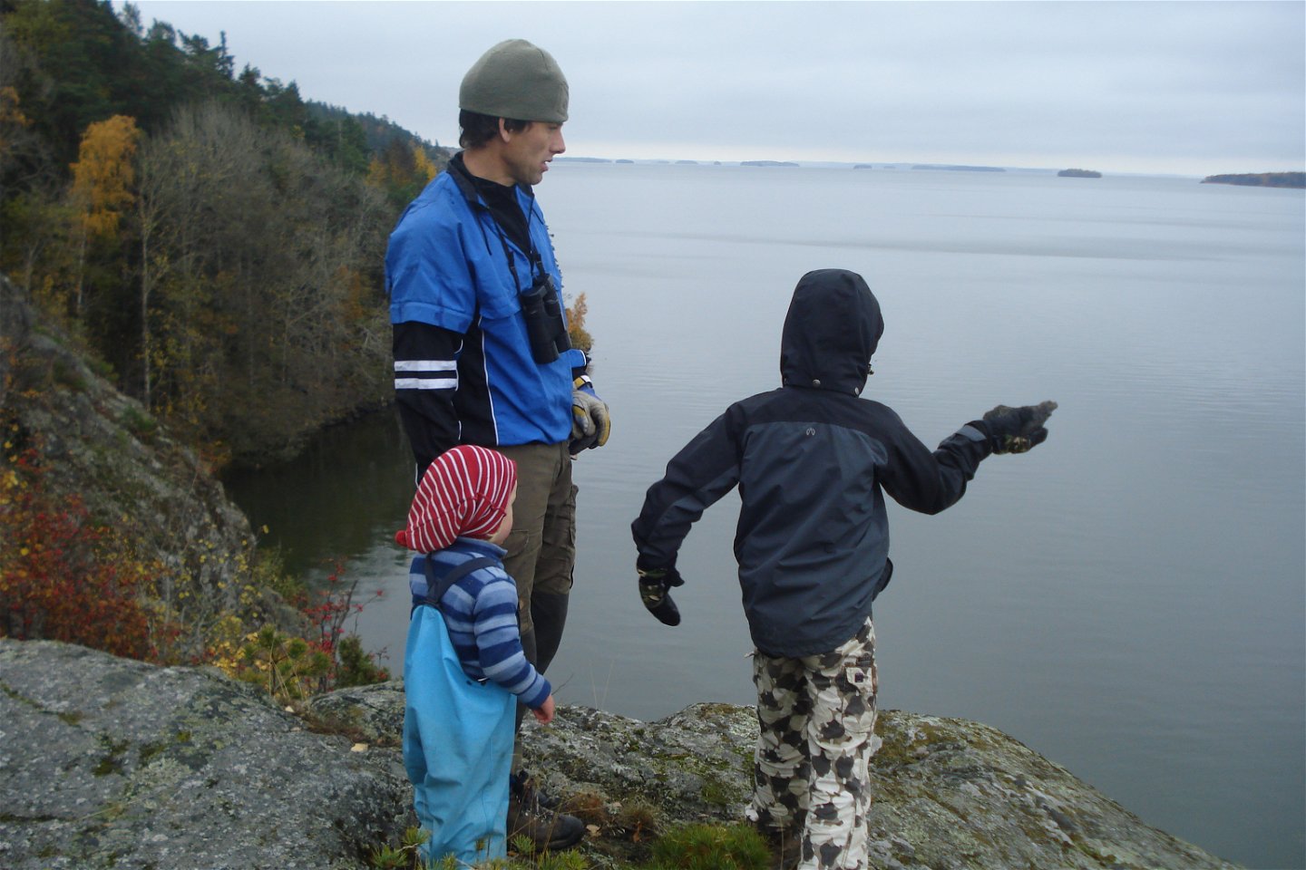 En man med ett större och ett mindre barn står vid en klippkant vid vattnet. 