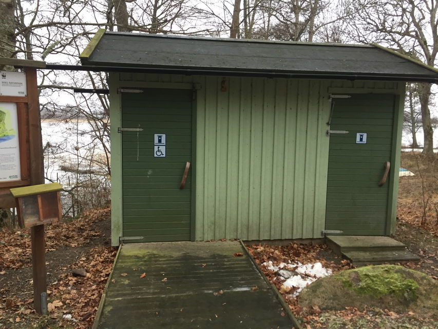 En torrtoalett med två dörrar står vid en strand. En ramp leder upp till ena dörren och en kort trappa leder upp till den andra.