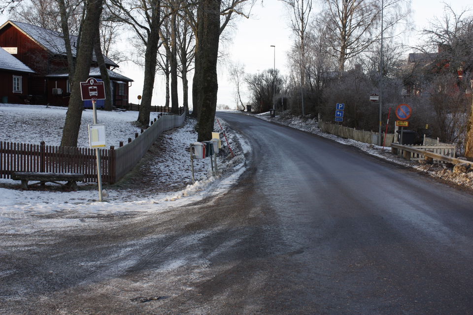 Vid sidan av en bilväg står en skylt för busshållplats. I bakgrunden står höga träd och ett hus.