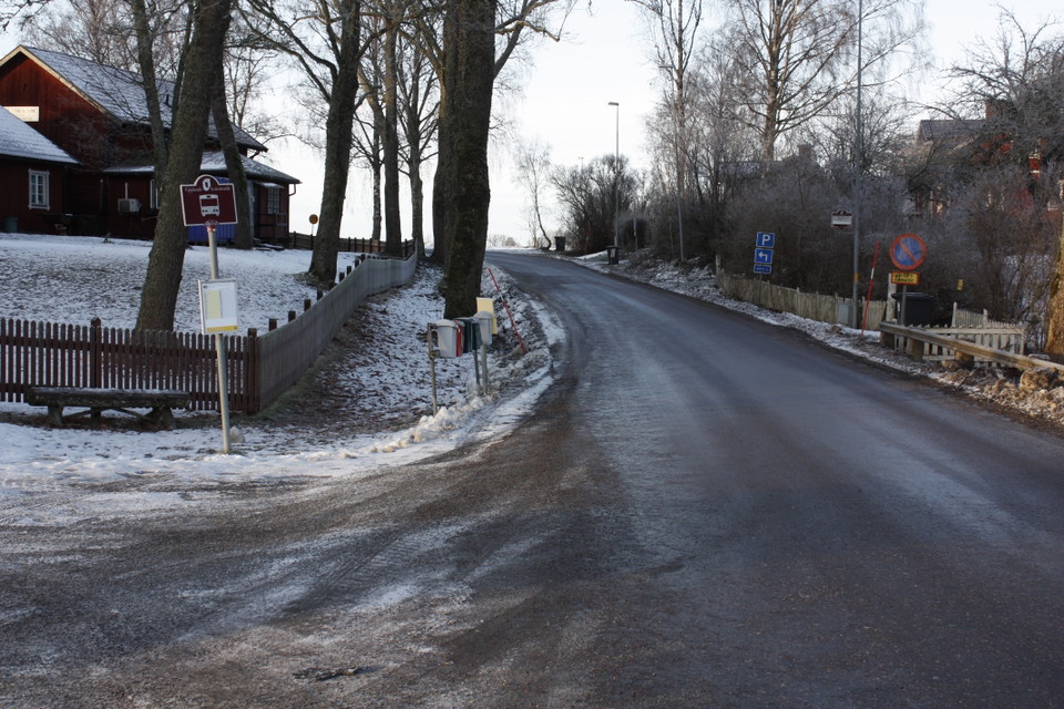 Vid sidan av en bilväg står en skylt för busshållplats. I bakgrunden står höga träd och ett hus.