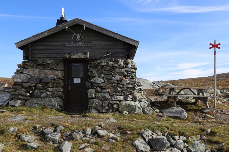 Ett stenhus med en bänk bredvid och ett rött ledkryss på ett fjäll