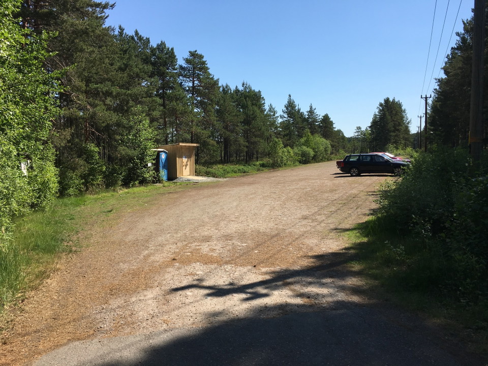 En stor parkeringsplats med några bilar i bortre hörnet. På parkeringen finns en tillgänglighetsanpassad torrtoalett och en utomhustoalett bredvid.