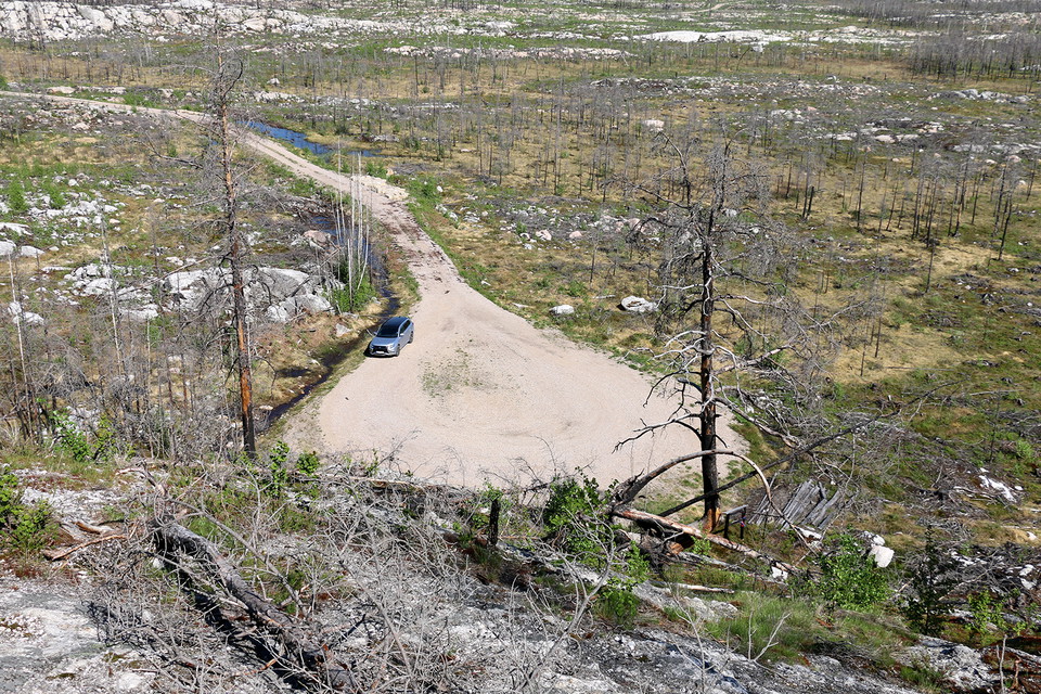 Vy ut över grusplan och bränt skogslandskap. En solverfärgad bil står parkerad på grusplanen.