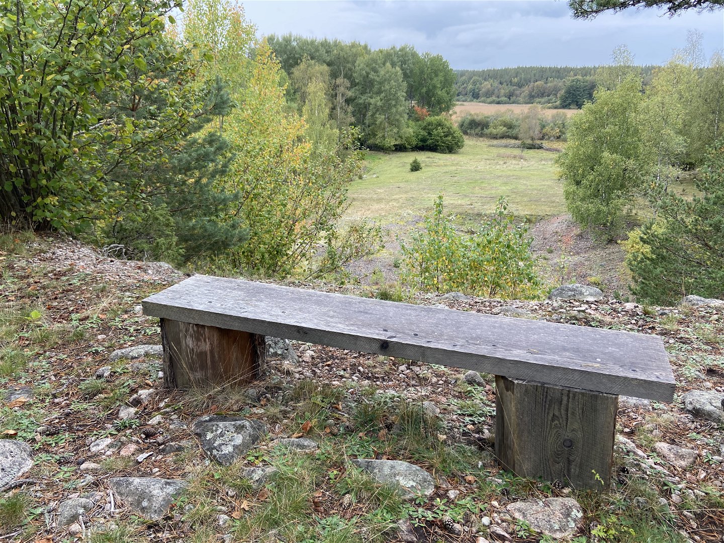 En sittbänk står på en stenig yta. I bakgrunden ser man vy över hagmark.