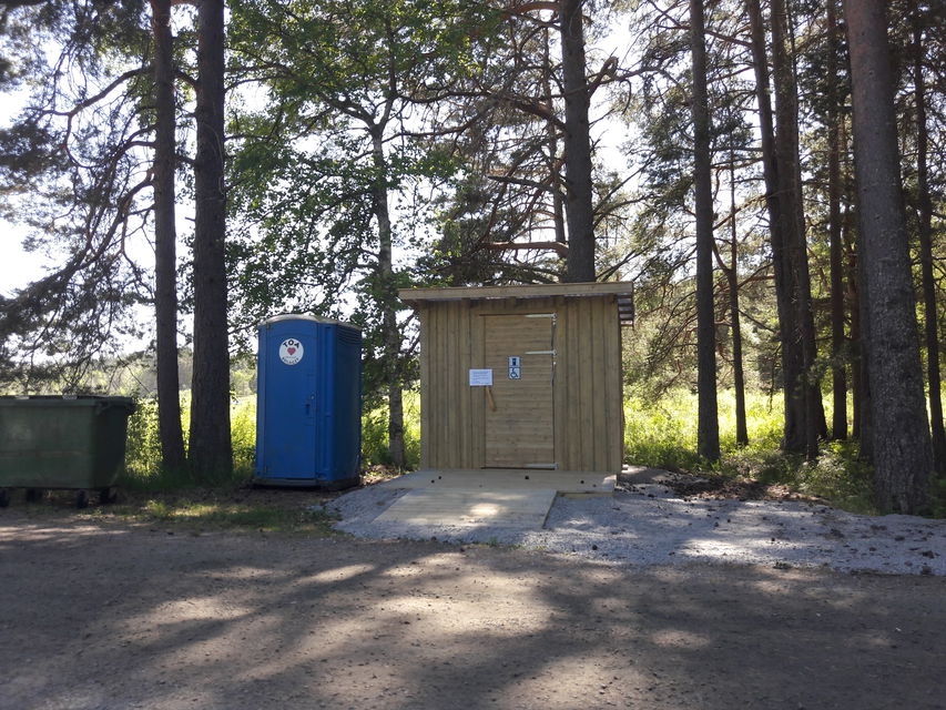 En ramp leder upp till en tillgänglighetsanpassad torrtoalett. Bredvid står en utomhustoalett och en soptunna.