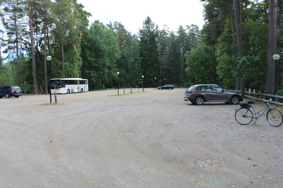 På en mycket rymlig grusad parkeringsplats står flera bilar, en buss och en cykel parkerade. Det finns belysning på parkeringen och den omges av träd.