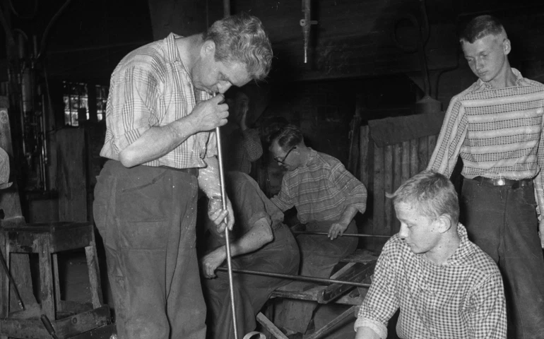 Två pojkar i glashyttan på Elme glasbruk studerar en äldre man som blåser glas. Mannen blåser ner i ett blåsrör riktat mot golvet. 