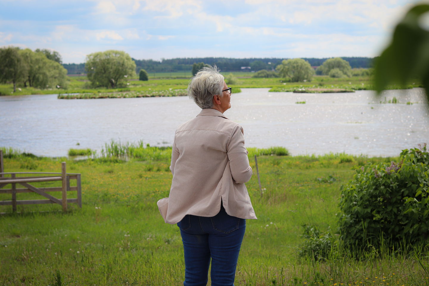 En äldre kvinna står och tittar ut över en liten sjö omgiven av grönt gräs.