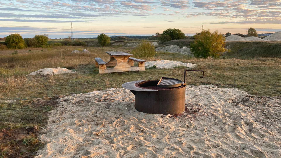 En rund grill omgiven av vit sand på marken. Bortom grillen tills ett rejält bänkbord i trä.