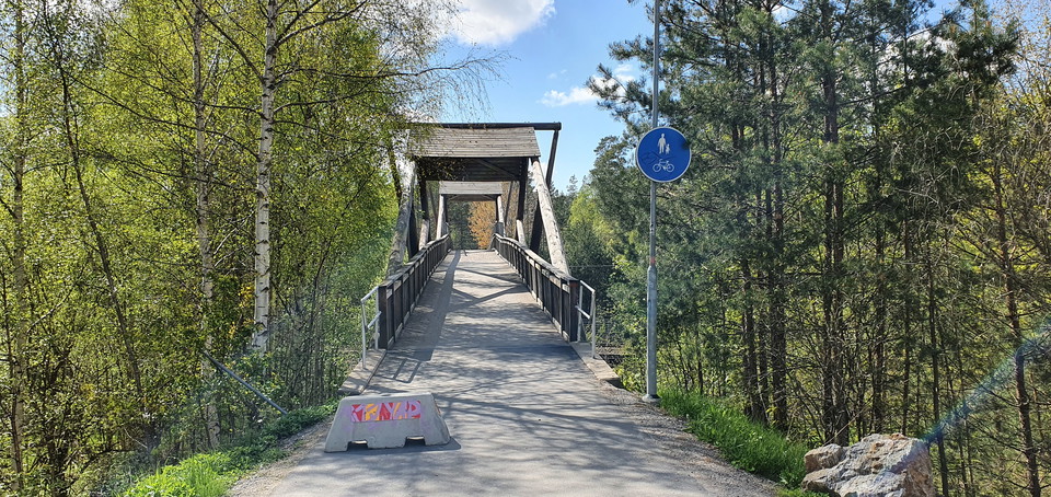 En asfalterad bro med räcken. Mitt på bron finns ett kort trätak. På vänster sida finns ett betonghinder.