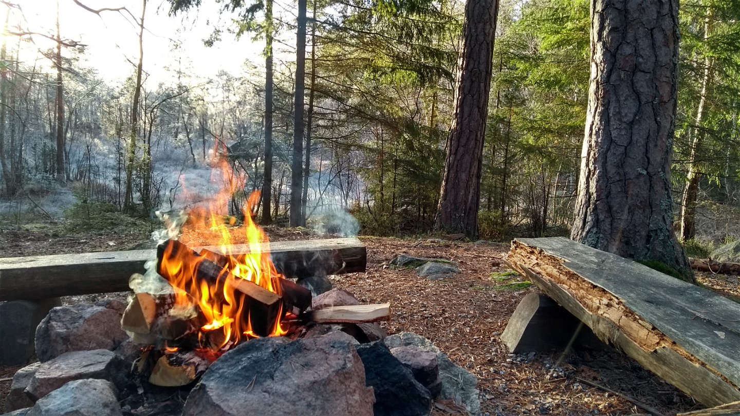 Närbild på en grillplats i skogen med sittbänkar runtom. På grillplatsen brinner en eld.