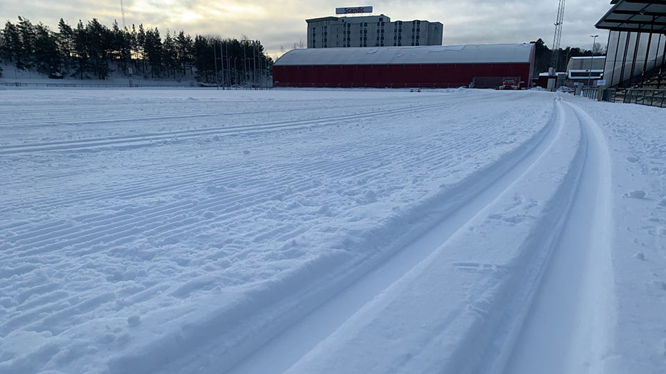 Närbild på skidspår i snö. I bakgrunden syns stora byggnader.