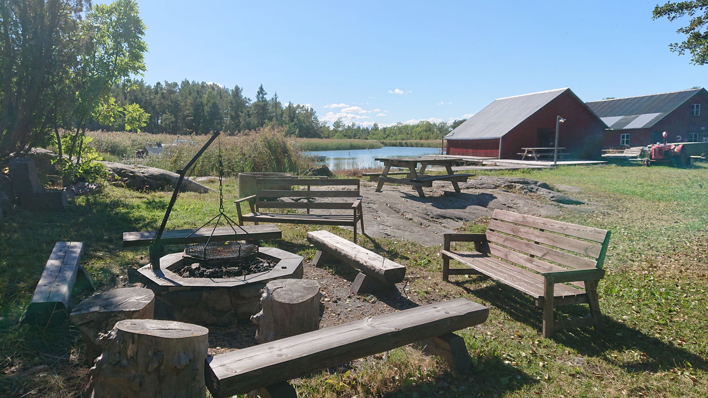 En låg grillplats med flera sittbänkar runtom står på gräsmark. I bakgrunden står ett bänkbord på en stenhäll och längre bort syns båthus och vatten.
