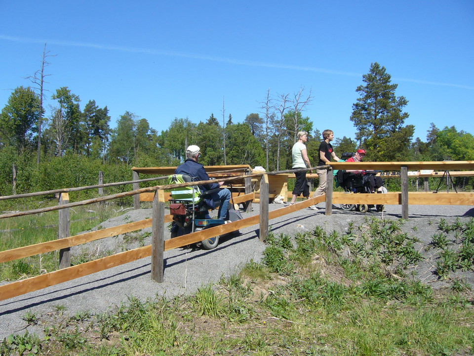 En person i permobil är på väg uppför en ramp som leder till en plattform. På plattformen finns en person i rullstol och två gående personer.