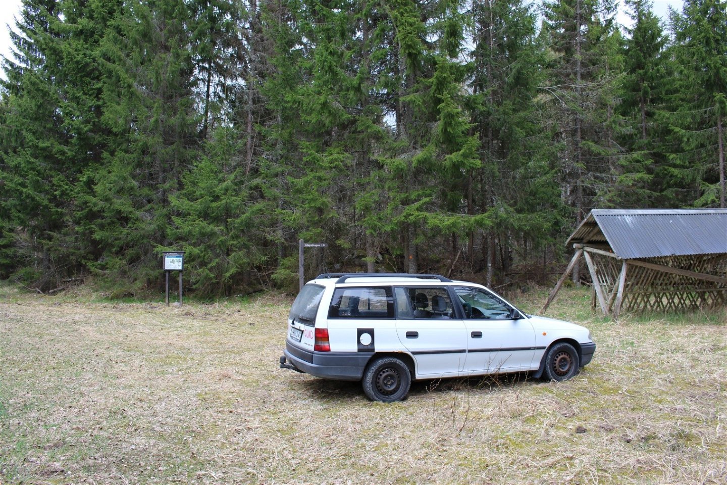 En bil står på något ojämn gräsmark. På platsen finns en informationsskylt och en vägvisare vid skogskanten.