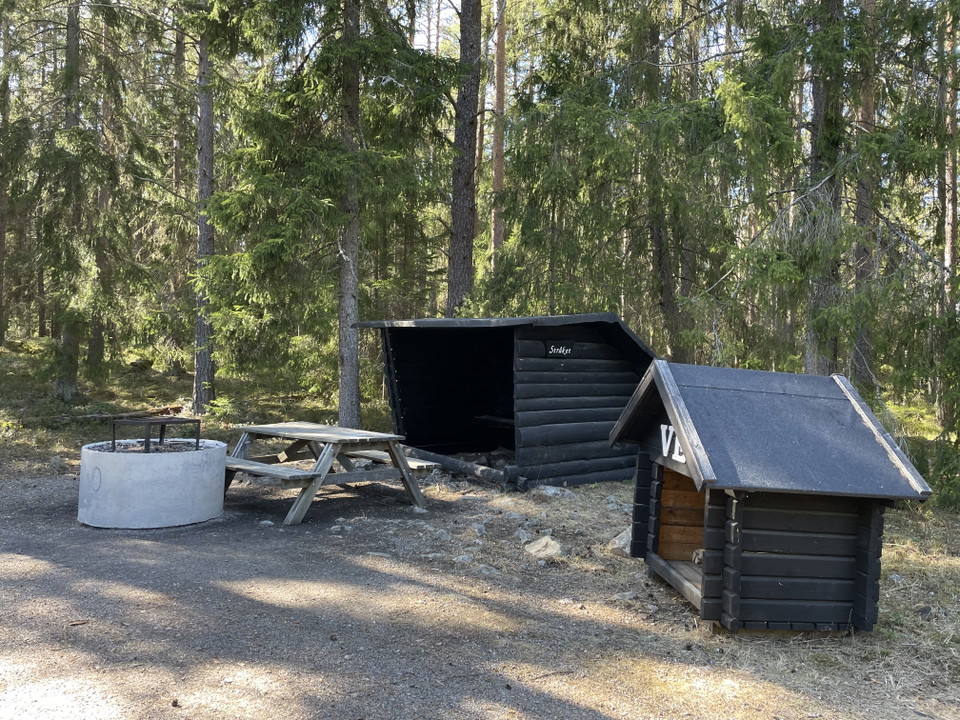 Rastplats vid namn stråket i skogsmiljö med ett svart vindskydd, eldstad, bänkbord och vedförråd