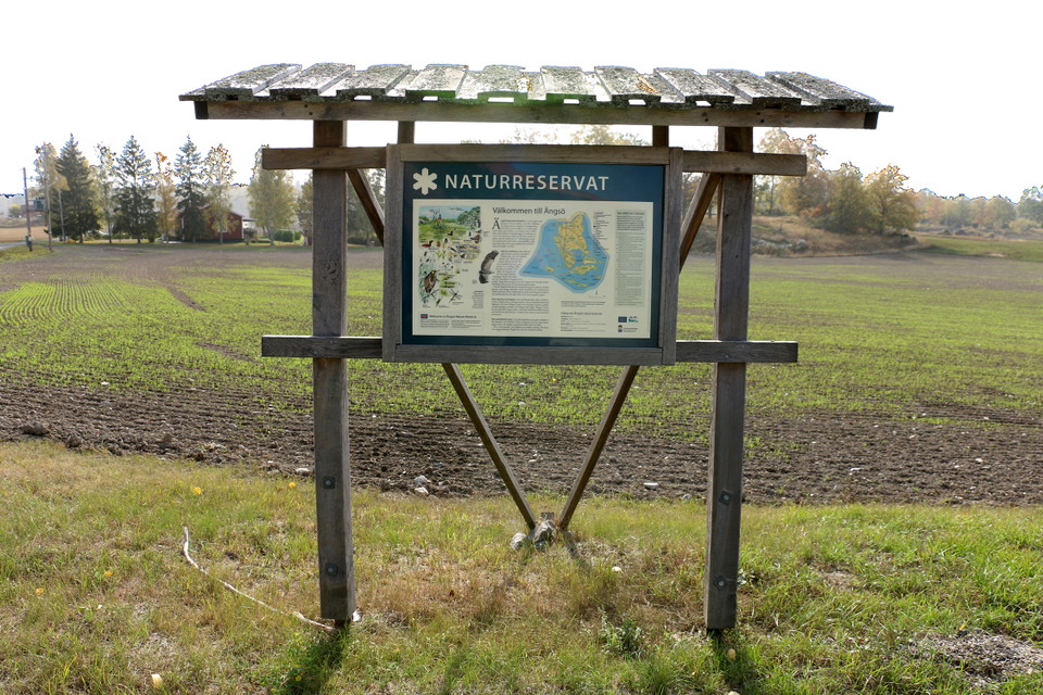 Informationstavla på en träställning. I bakgrunden ett åkerlandskap.