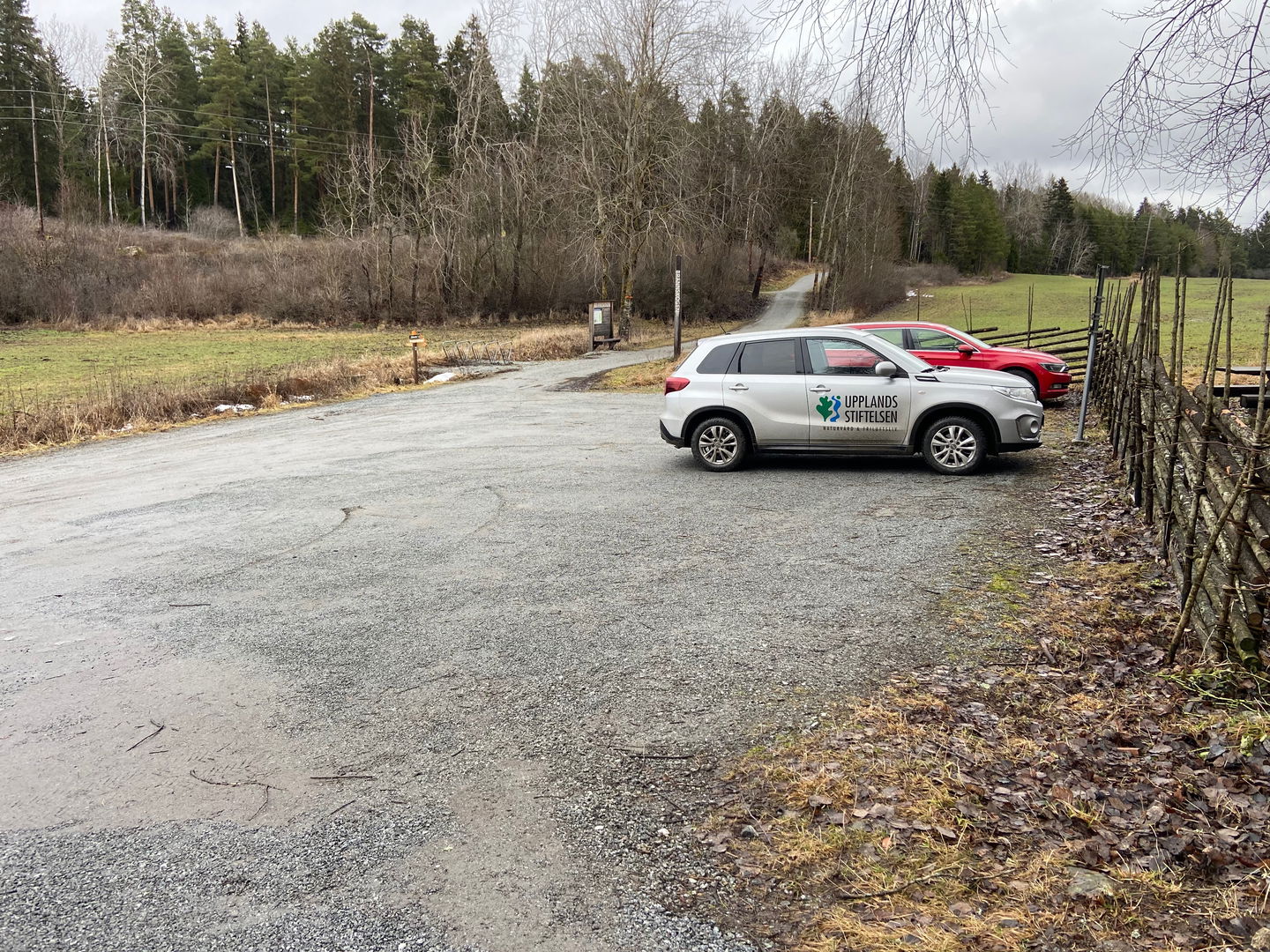 En grå och en röd bil står parkerade på en grusad yta. Framför bilarna syns en gärdesgård. I bakgrunden syns ett elljuspår, skog och hagmark.