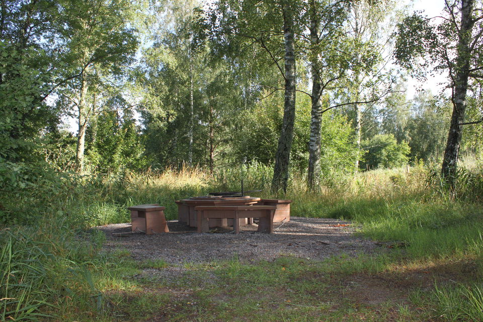 En grillplats med träbänkar, eldplats med grillgaller. Runt omkring syns träd och gräs.