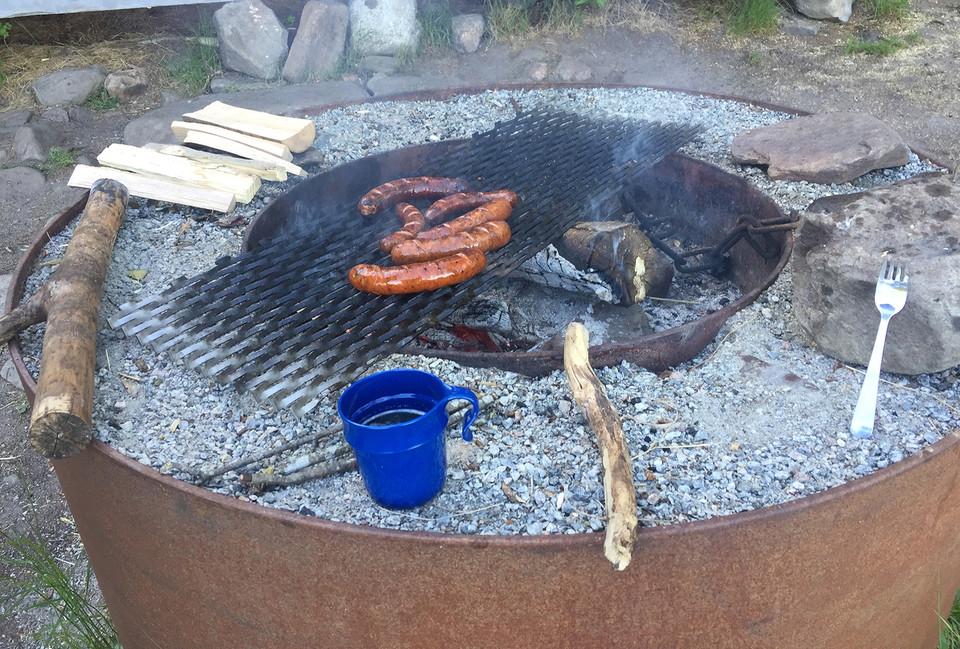 Sex korvar på ett galler över glöd. Vid grillen står en blå plastmugg och det ligger bestick på vardera sida av muggen.