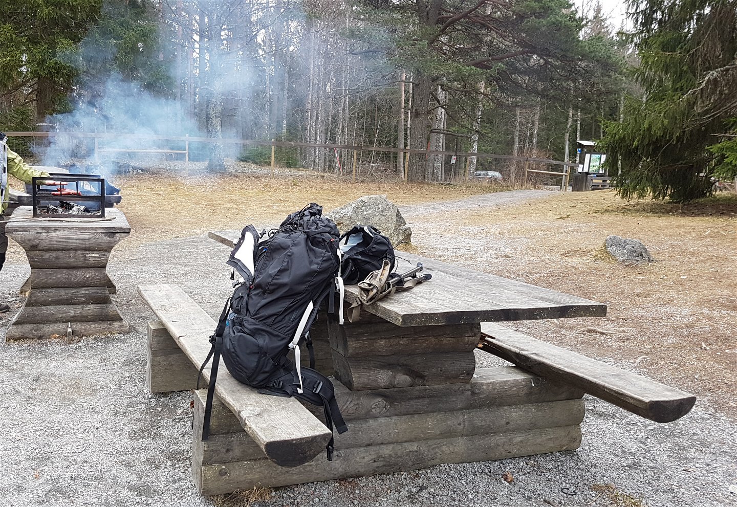 En ryggsäck ligger på ett bänkbord i skogen. I bakgrunden står en person vid ett grillbord och grillar.