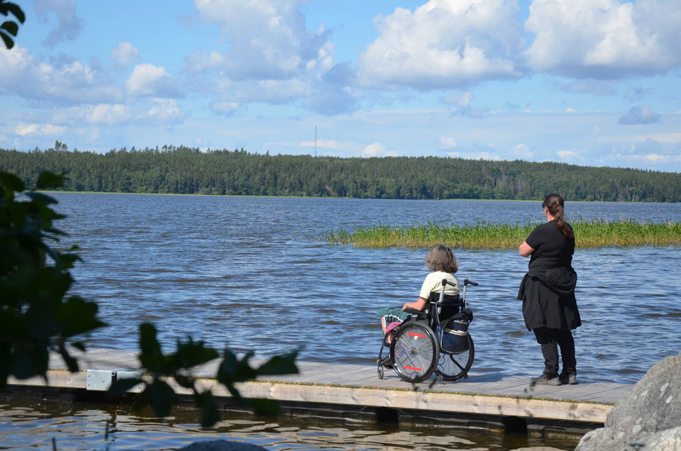 En stående kvinna och en kvinna i rullstol befinner sig på en brygga i trä som sträcker sig ut i vattnet.