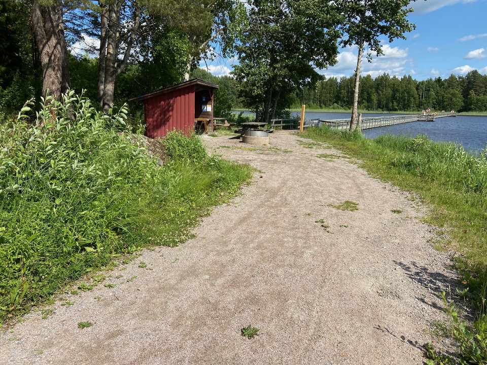 Sommarbild. Hårdgjord stig i förgrunden, skymtar rastplats och vatten