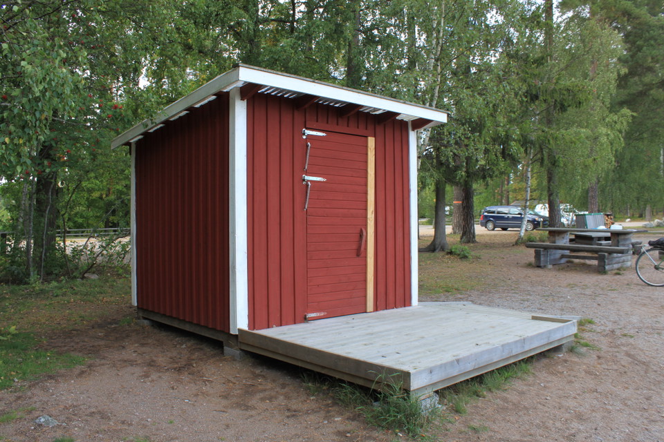 En torrtoalett med en träplattform framför. För att komma upp på plattformen måste man ta en kliv uppåt. I bakgrunden syns en grillplats och parkering.