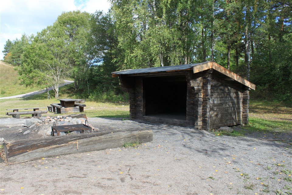 På en öppen yta står en låg grillplats med stockar runtom och två bänkbord bakom. Bredvid står ett vindskydd med lågt tak och en tröskel in i vindskyddet.
