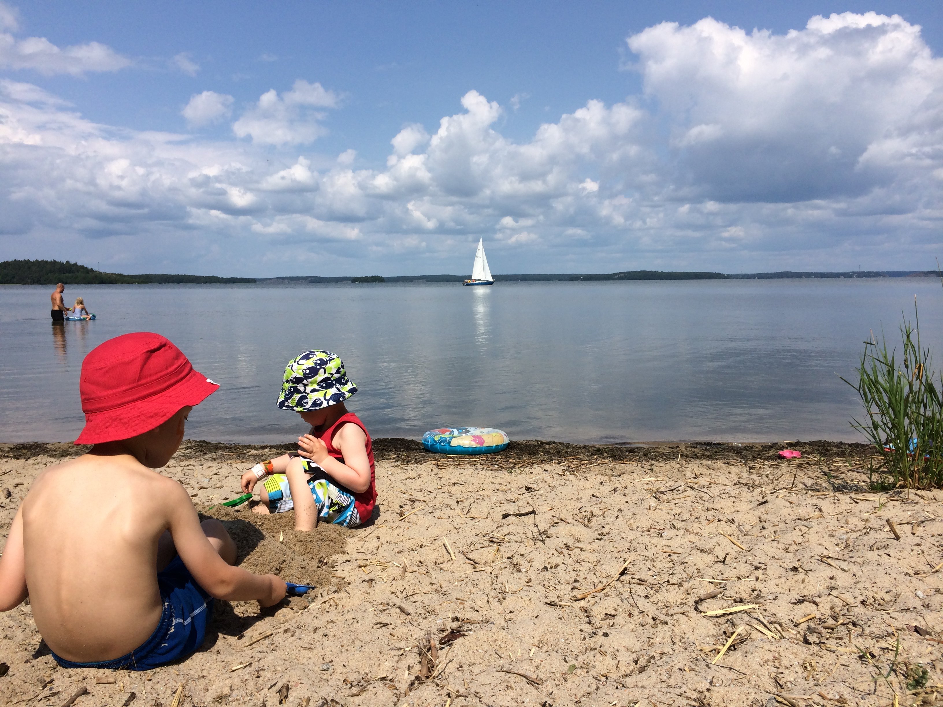 Två barn leker på en sandstrand. I vattnet badar två personer och en segelbåt syns längre ut. 