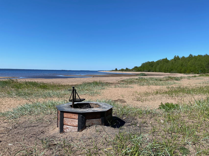 På en strand med sand och vass står en grillplats med vridbart grillgaller
