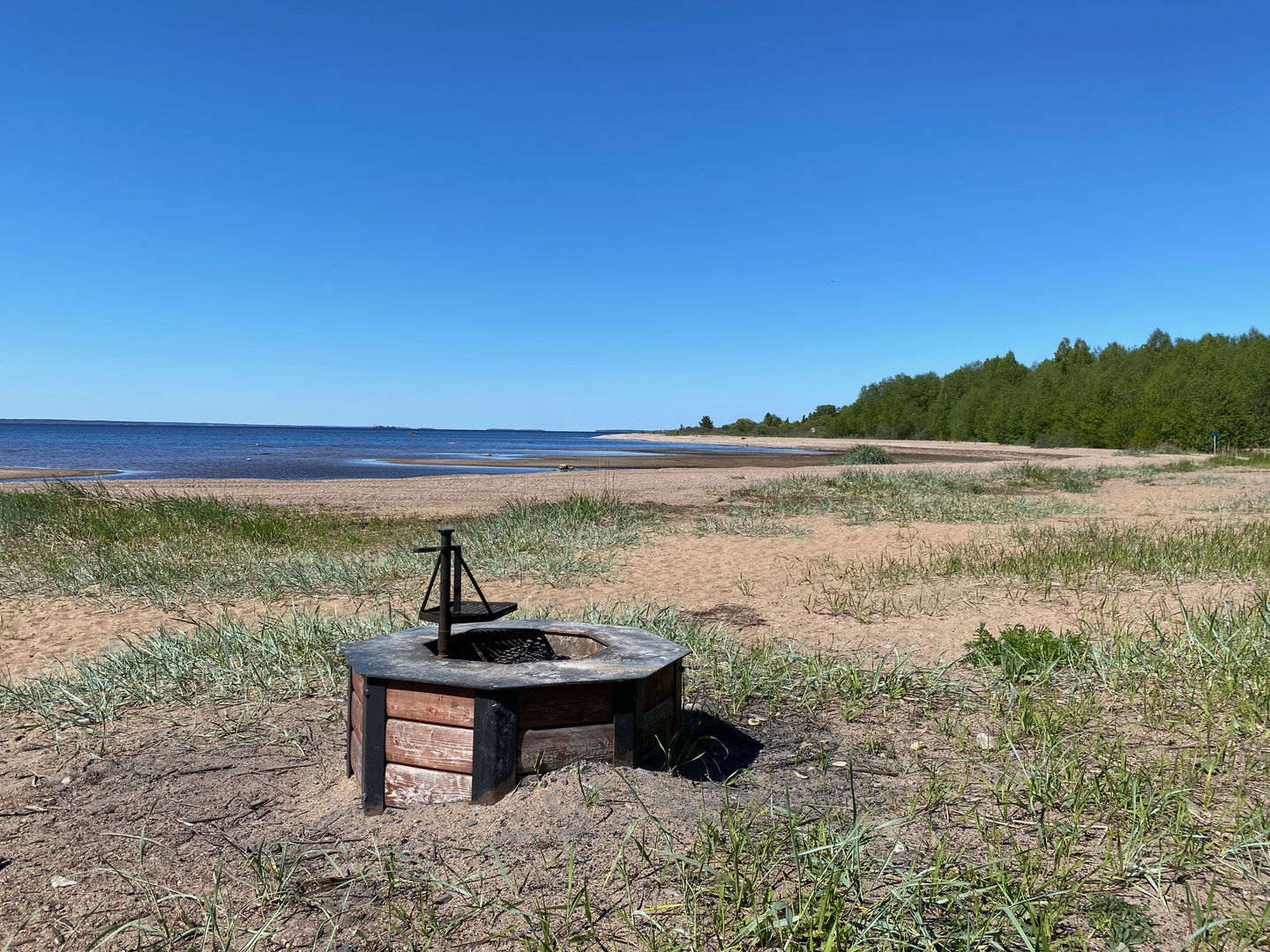 På en strand med sand och vass står en grillplats med vridbart grillgaller